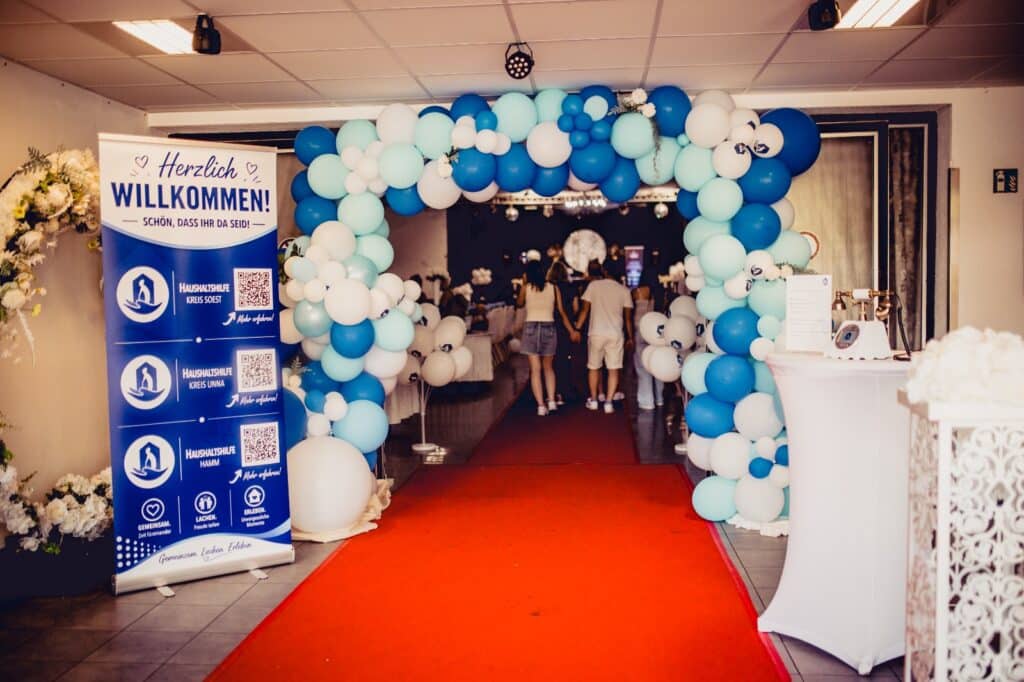 Sommerfest der Haushaltshilfe Kreis Soest: Empfang mit blau-weißem Ballonbogen, rotem Teppich und Willkommensbanner in der Bördelandhalle Welver